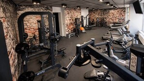 Ein Fitnessraum mit unterschiedlichen Trainingsgeräten, einer Ziegelwand und einer Decke mit Rasterleuchten.