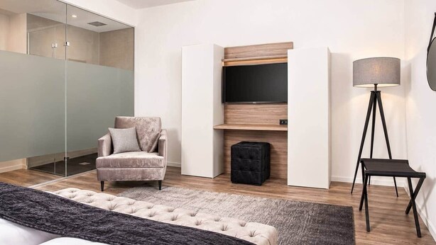 Ein modernes Hotelzimmer mit einem Bett, einer Sitzecke mit einem Sessel, einem kleinen Hocker, einer Stehlampe und einem an einer Holzvertäfelung befestigten Fernseher.