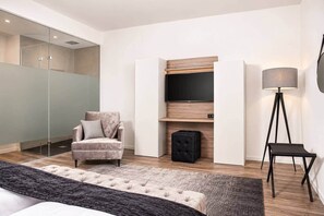 A modern hotel room with a bed, a seating area with a chair, a small ottoman, a floor lamp, and a television mounted on a wooden panel. - The Faces | Suite | Balcony | 001 (Berlin)