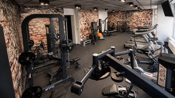 A gym with various exercise equipment, a brick wall, and a ceiling with grid lights. - The Faces | Premium Studio | Balcony | 204 (Berlin)