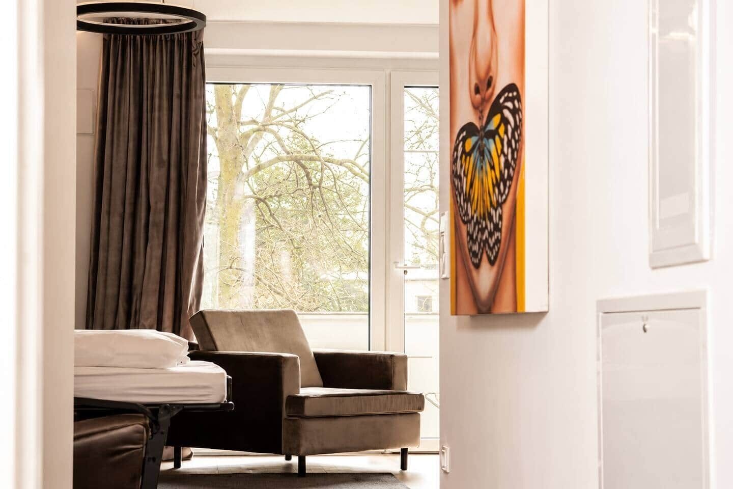 Une chambre d’hôtel avec un lit, une chaise, une fenêtre avec des rideaux et un tableau au mur.