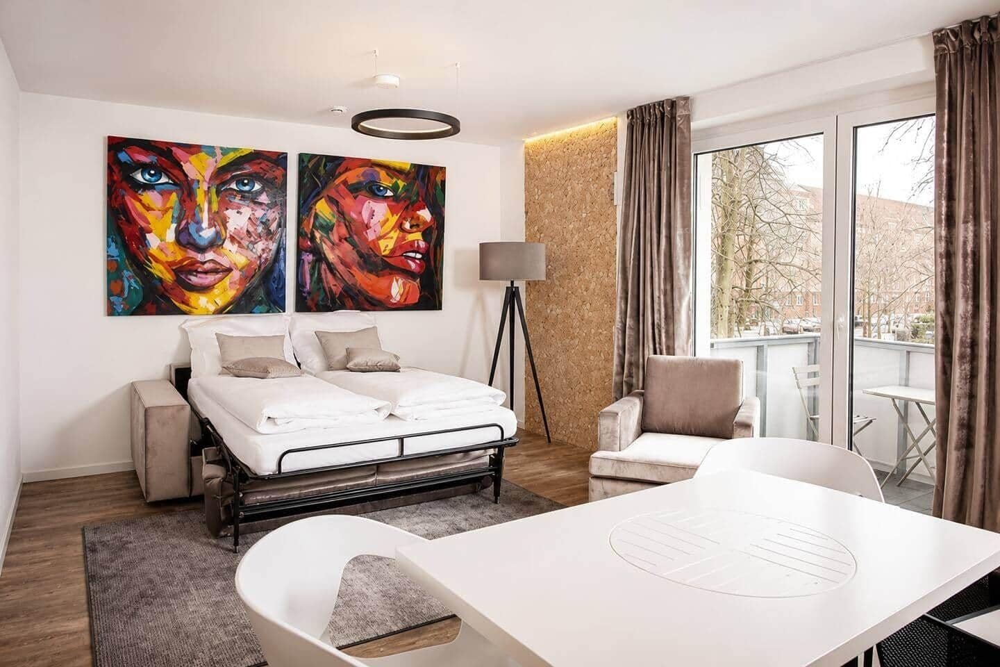 A modern hotel room with a bed, a desk, a chair, a lamp, and two large paintings on the wall.