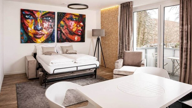 Et moderne hotelværelse med seng, skrivebord, stol, lampe og to store billeder på væggen.