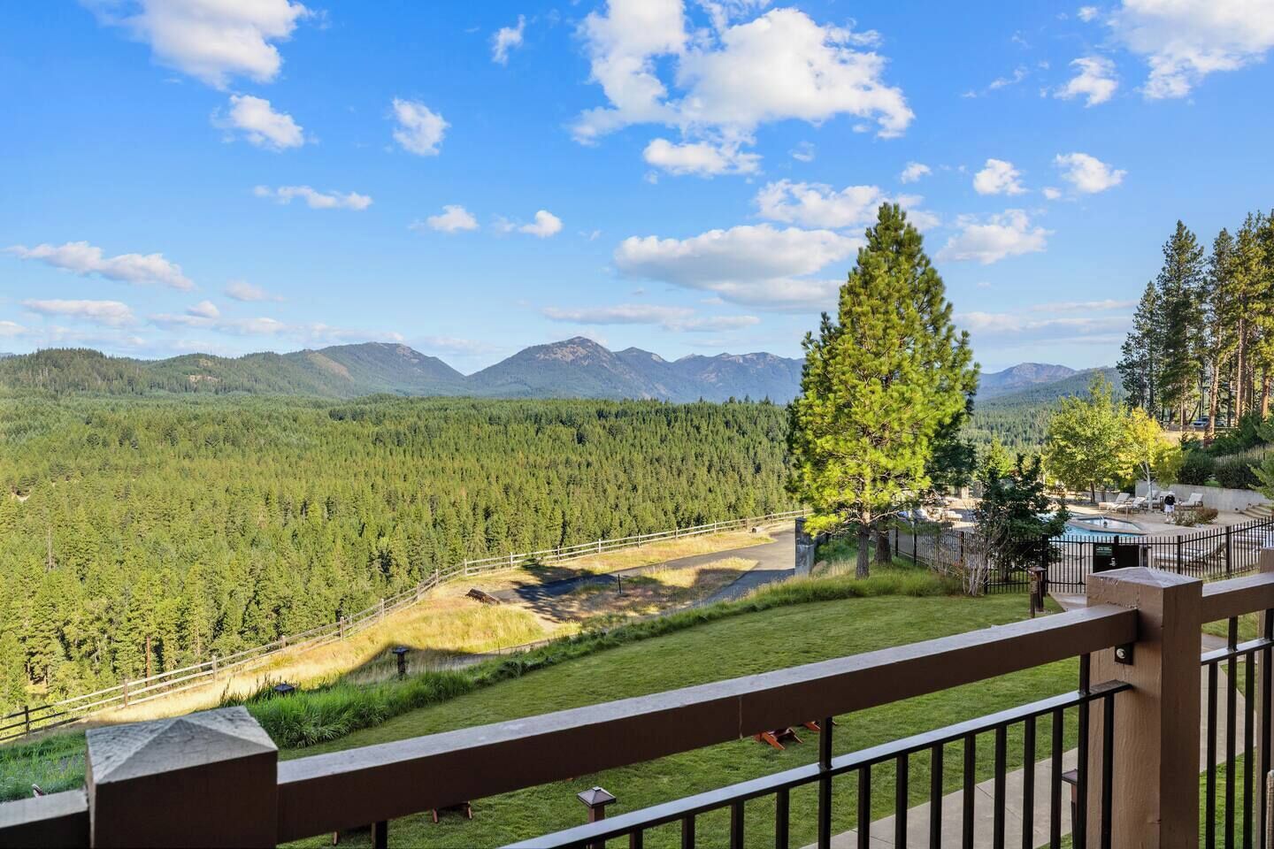 A view from a balcony with a pool, surrounded by a forest and mountains.