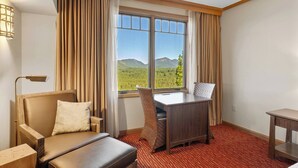 A hotel room with a bed, a desk, a chair, and a view of a forest and mountains.