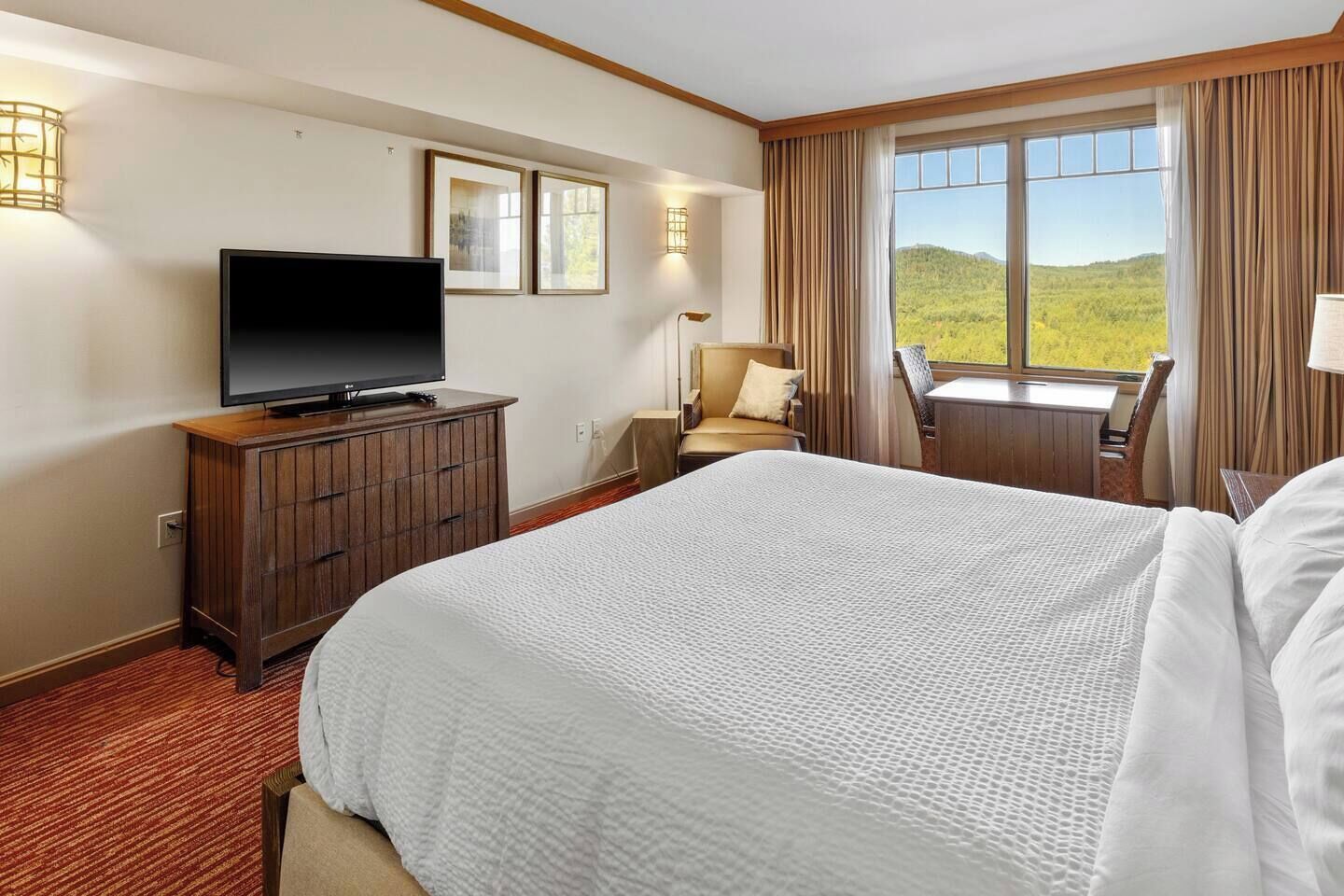 A hotel room with a large bed, a television on a wooden cabinet, a desk with a chair, and a view of a green landscape through the window.