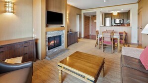 A modern living room with a fireplace, a flat-screen TV, a dining area with a table and chairs, and a kitchen in the background.
