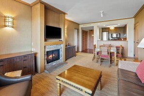 A modern living room with a fireplace, a flat-screen TV, a dining area with a table and chairs, and a kitchen in the background.