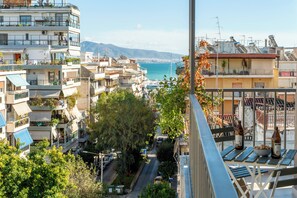A residential area with multiple-story buildings, a street with parked cars, and a view of the sea in the background. - Pame Rentals – Olive Executive Suite D3 (Pireas)