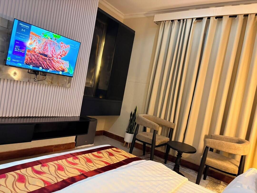 A hotel room with a bed, a flat-screen TV mounted on the wall, a window with curtains, and a small table with a chair.