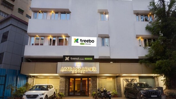 A modern building with a sign for 'treebo Ambassador Hotel' and a parking area with vehicles. - Treebo Trend Ambassador (Ahmedabad)