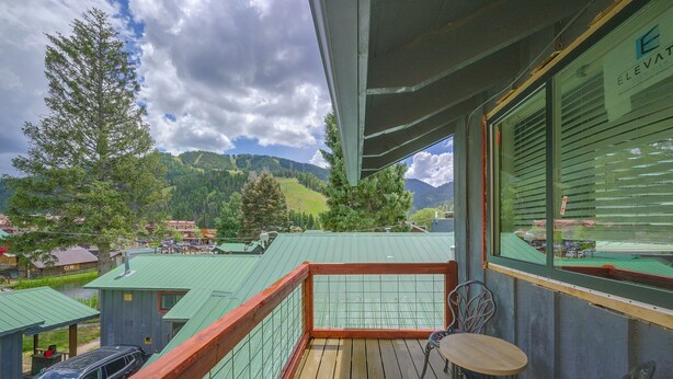 A balcony with a wooden floor and metal railing, overlooking a mountainous landscape with green fields and buildings. - Walk to Ski Lifts! Apt in Heart of Red River (Red River)