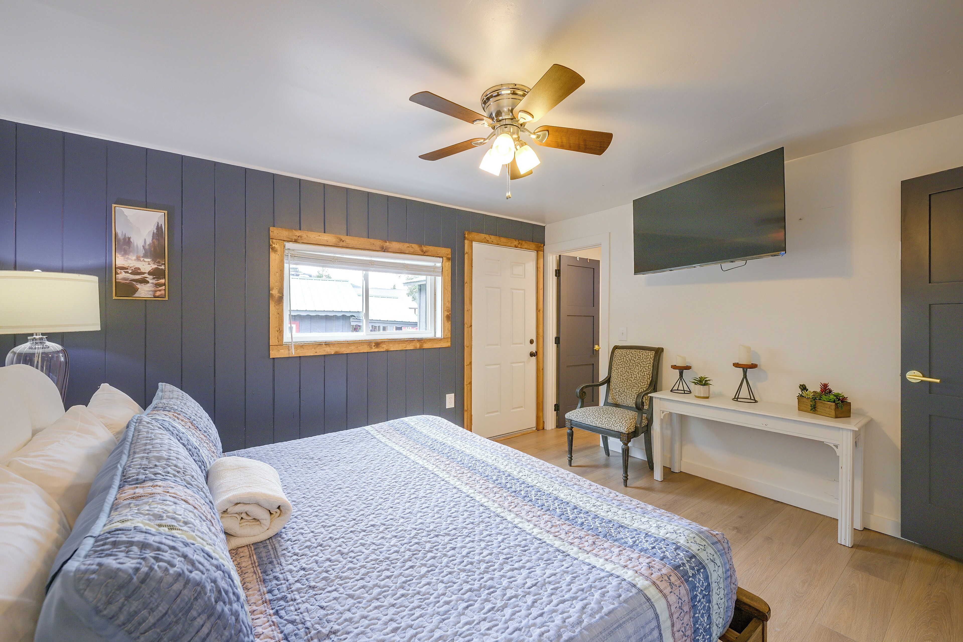 A bedroom with a bed, a chair, a TV, a ceiling fan, and a window.