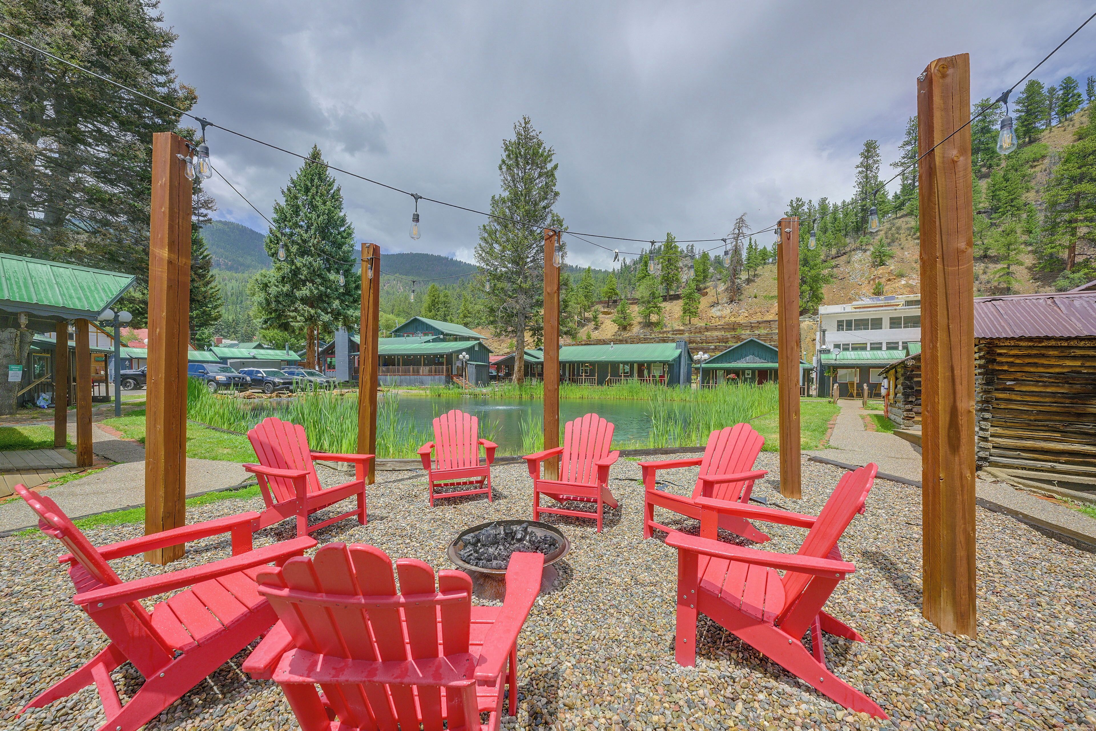 A seating area with red chairs and a fire pit, surrounded by a gravel path and greenery, with buildings and a lake in the background.