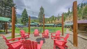 A seating area with red chairs and a fire pit, surrounded by a gravel path and greenery, with buildings and a lake in the background. - Walk to Main St: Red River Lodge w/ Ski Views! (Red River)