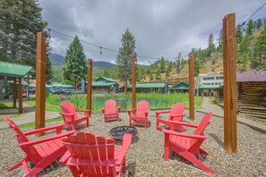A seating area with red chairs and a fire pit, surrounded by a gravel path and greenery, with buildings and a lake in the background. - Walk to Main St: Red River Lodge w/ Ski Views! (Red River)