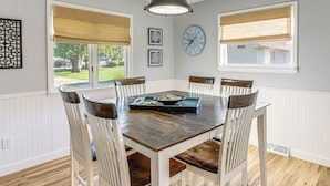Un comedor con una mesa de madera y sillas blancas, un reloj en la pared y ventanas con persianas.
