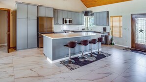 A modern kitchen with grey cabinets, a stainless steel refrigerator, a microwave, and a large island with bar stools. - Gated Ranch Retreat Near Lakes & Frisco (Aubrey)