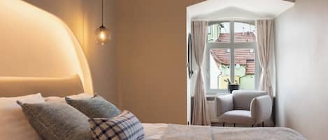 Un dormitorio con cama, almohadas, una silla y una ventana con cortinas.