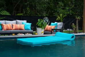 A pool area with a blue floating mat, a gray sofa with colorful cushions, and a black fence with greenery in the background. - Hidden Sanctuary with Heated Saltwater pool, fire pit, and koi pond (St. Augustine)