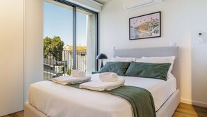 Una habitación de hotel con cama, ventana con vista a edificios, un cuadro en la pared y un termostato.