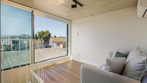Una sala de estar moderna con un amplio ventanal corredero que da a un balcón, un sofá gris con cojines y piso de madera.