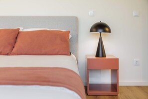 Ein ordentlich bezogenes Bett mit grauem Kopfteil und zwei orangefarbenen Kissen, eine schwarze Lampe auf einem pinkfarbenen Nachttisch und eine weiße Wand mit zwei Lichtschaltern.