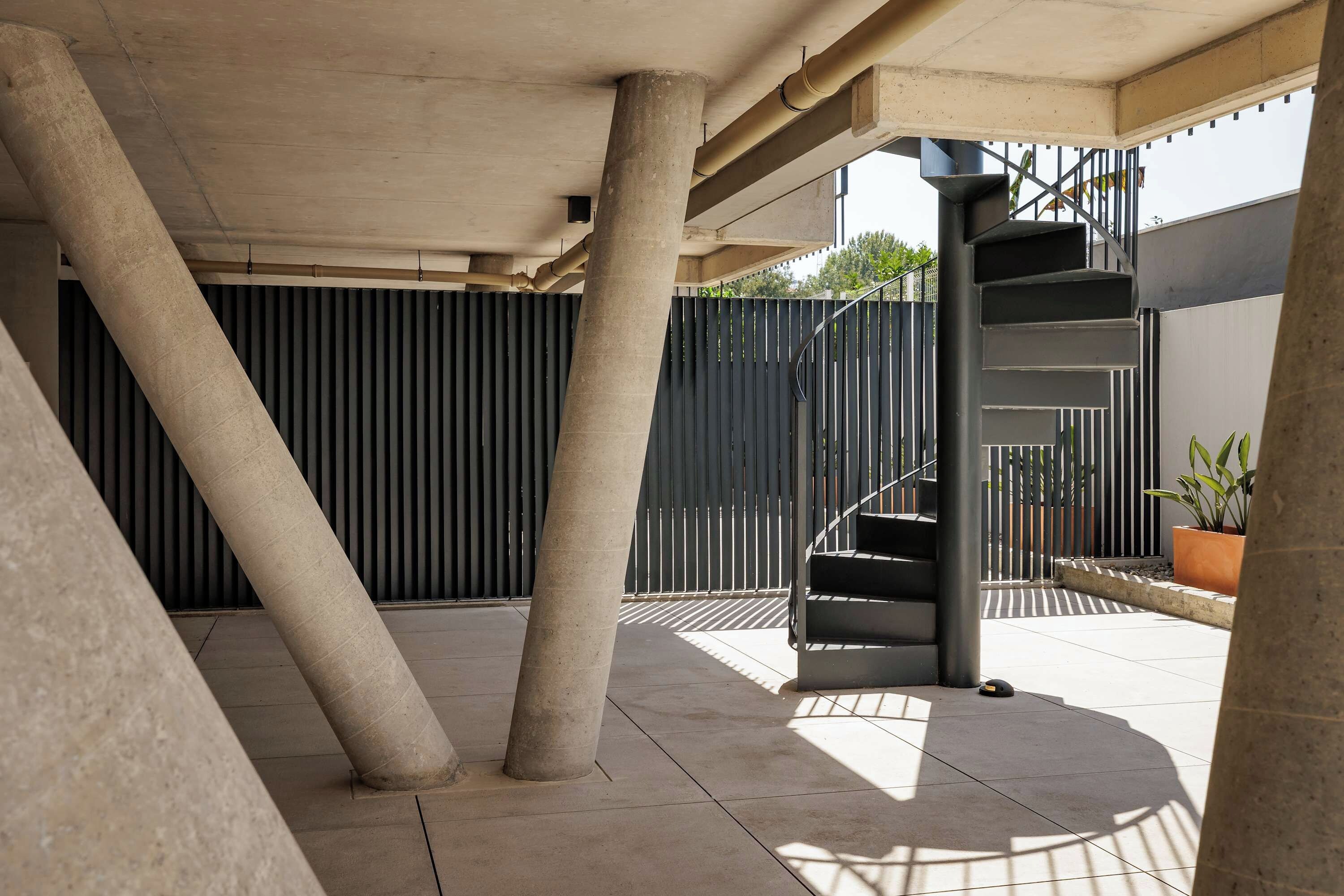 Un espace extérieur abrité, doté de piliers en béton, d’un escalier en métal et d’une clôture noire.