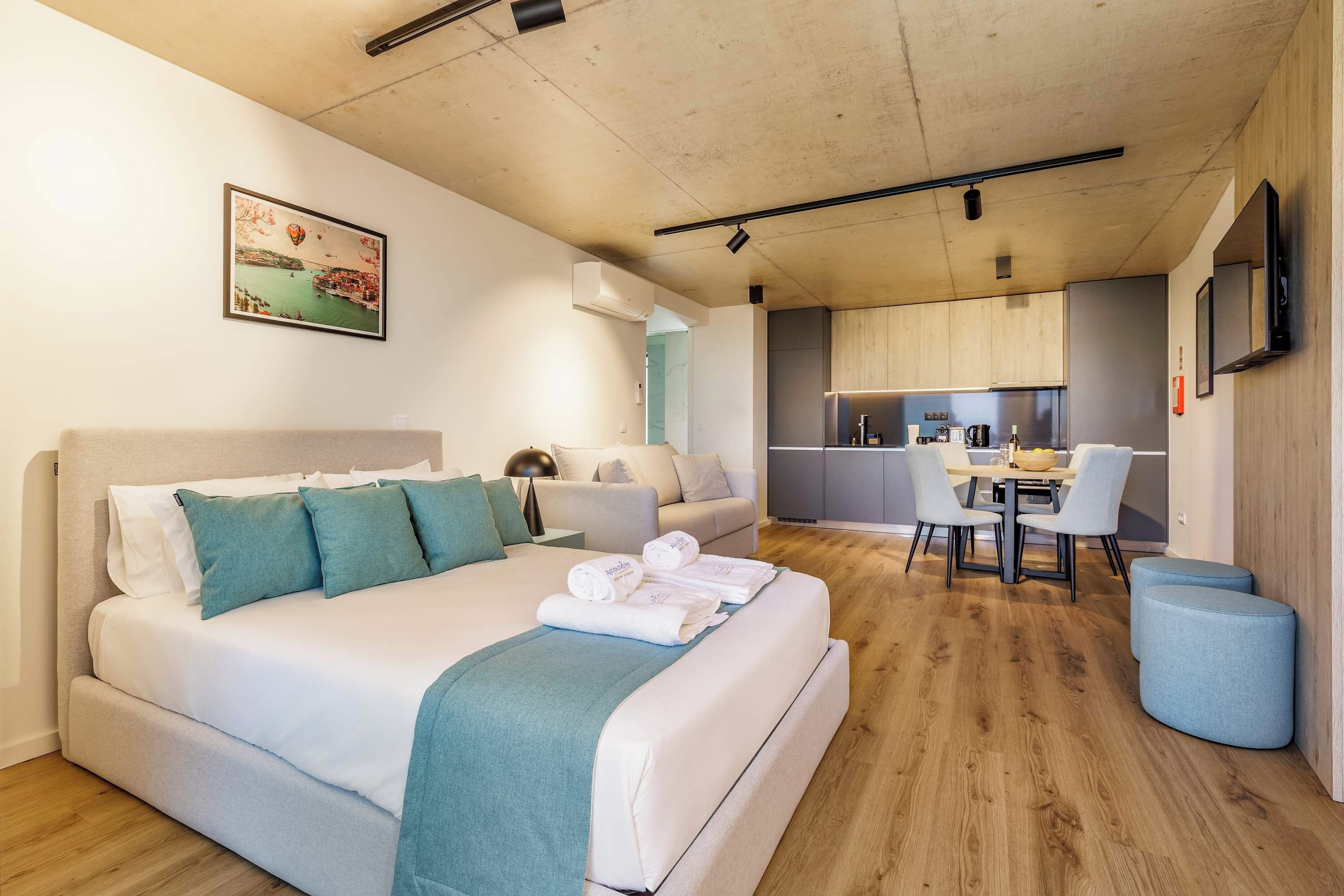A modern hotel room with a bed, a sofa, a dining table with chairs, a kitchen area, and a mounted TV.