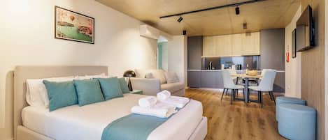 A modern hotel room with a bed, a sofa, a dining table with chairs, a kitchen area, and a mounted TV.