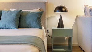 En pænt redt seng med gråt sengegavl, et lille natbord med lampe og en stikkontakt.
