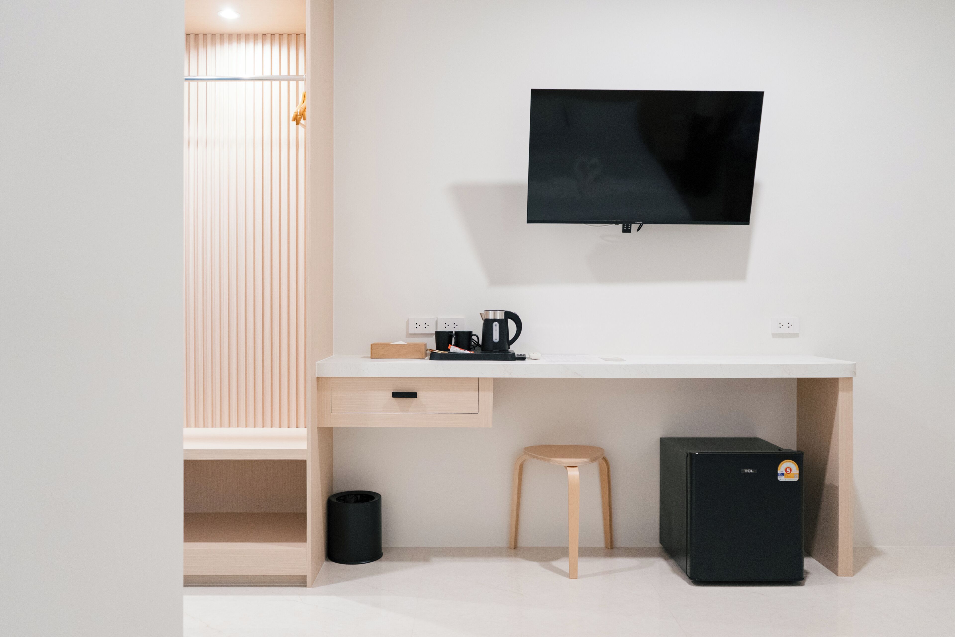 Una habitación minimalista con un escritorio blanco, un televisor negro montado en la pared, un taburete pequeño y un mueble negro para almacenamiento.
