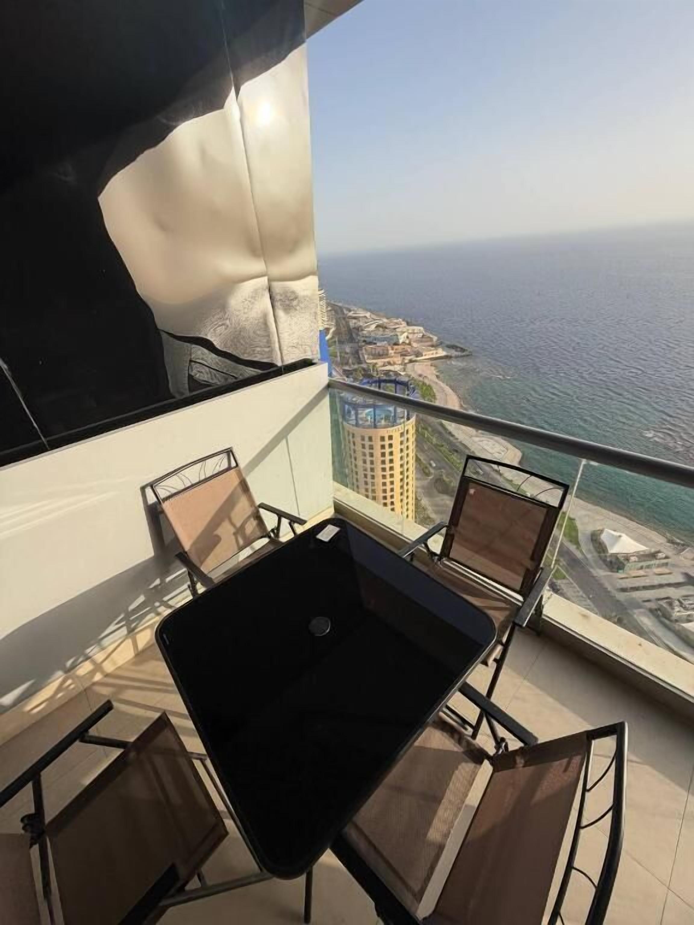 A balcony with a table and chairs, overlooking a coastal cityscape with buildings and a body of water.