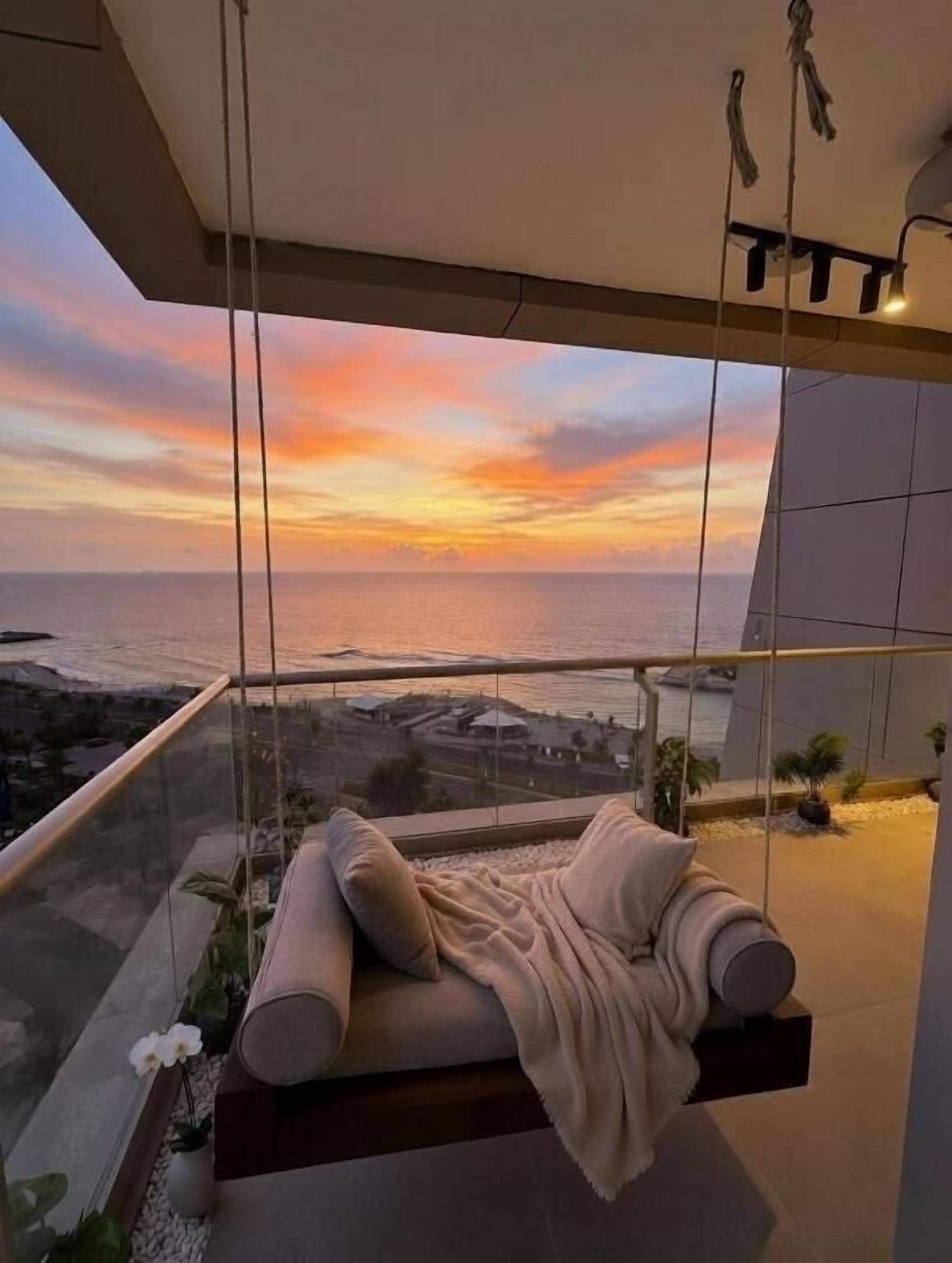 Un balcón con vista al océano al atardecer, un área de descanso con cojines y una lámpara colgante.