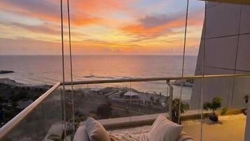 Un balcón con vista al océano al atardecer, un área de descanso con cojines y una lámpara colgante.