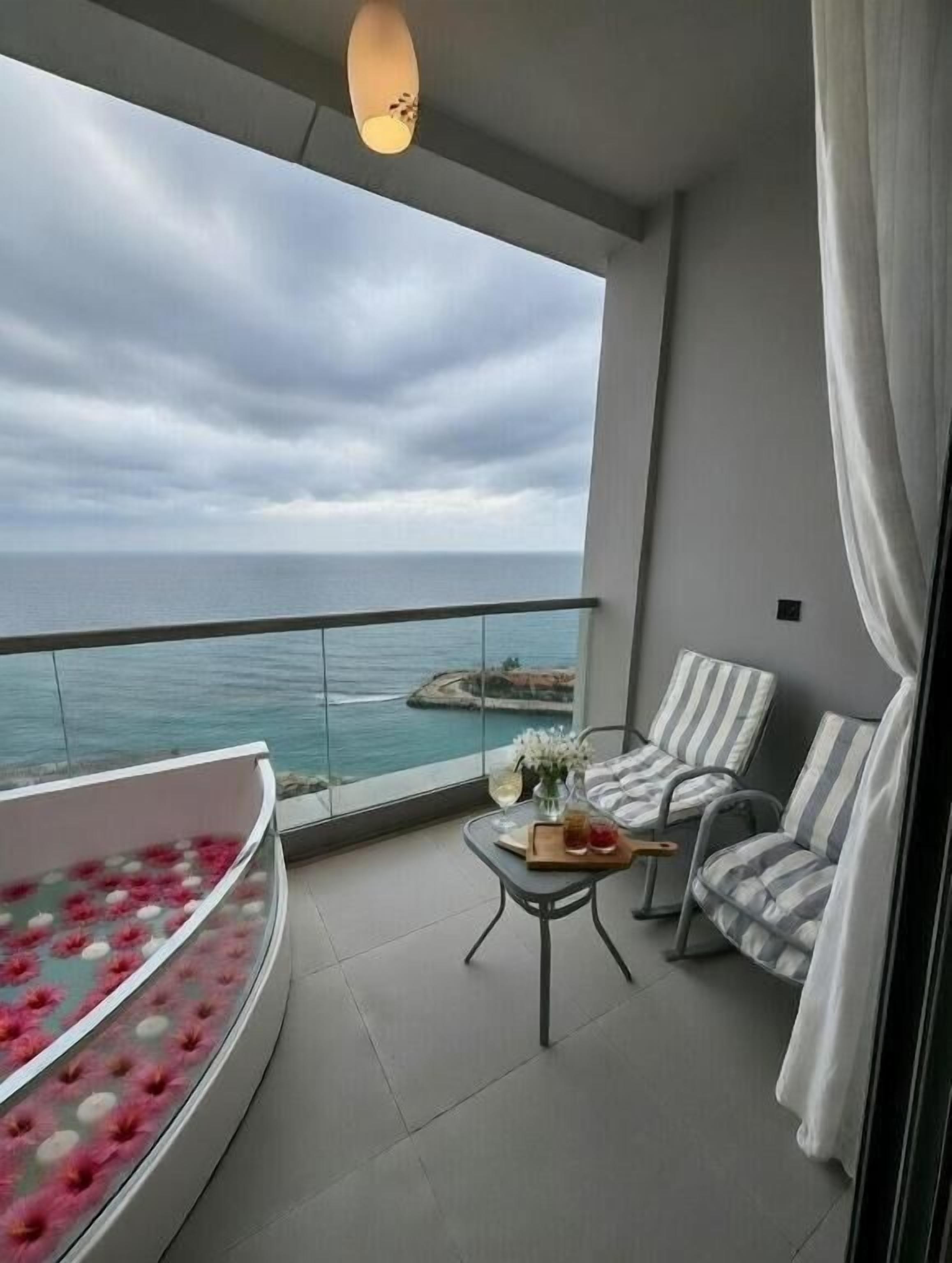 Un balcon avec une baignoire à bords arrondis, une table avec une boisson et des chaises à rayures.