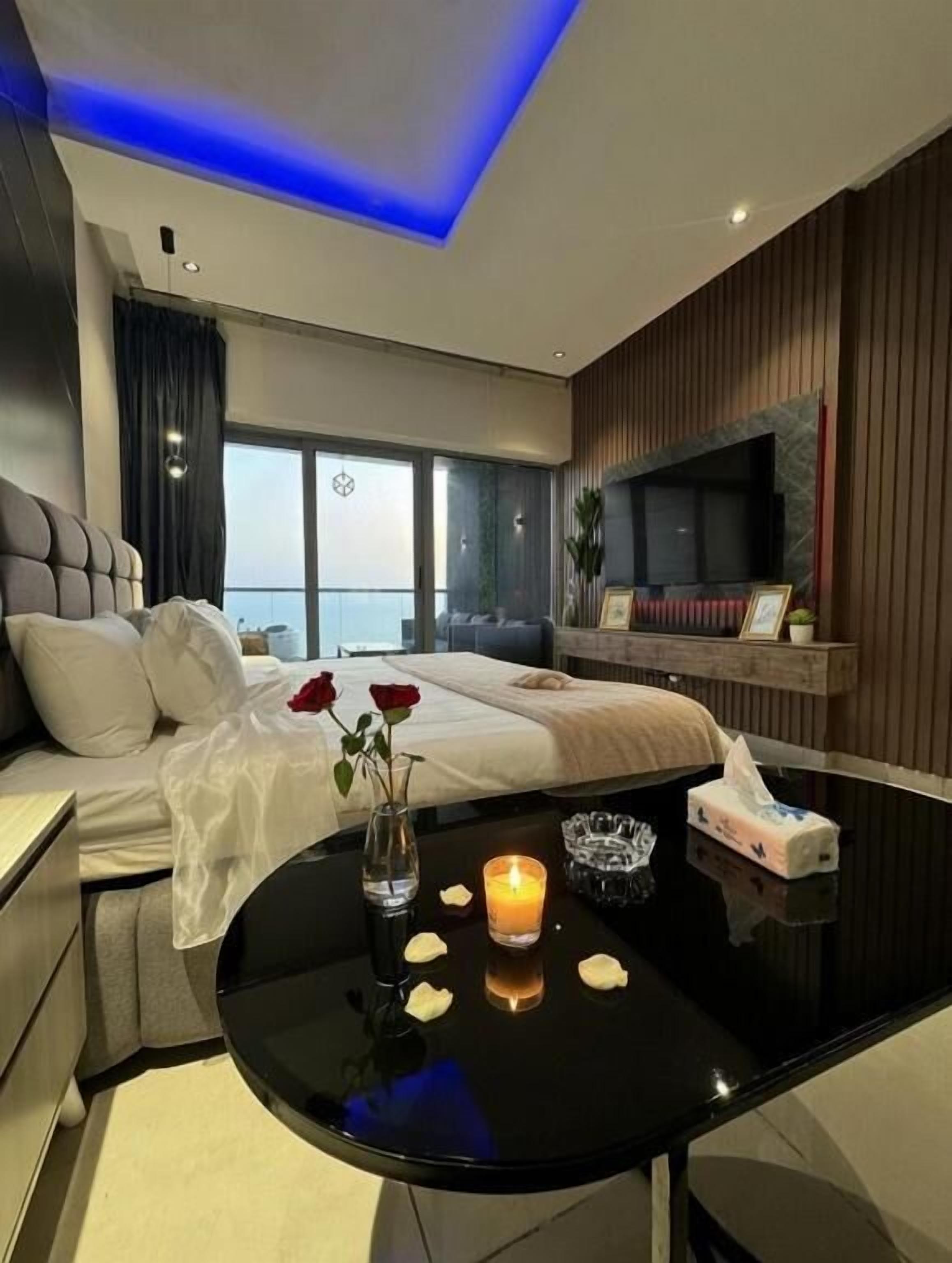 A modern hotel room with a bed, a flat-screen TV, a round black table with a candle, and a view of the city.