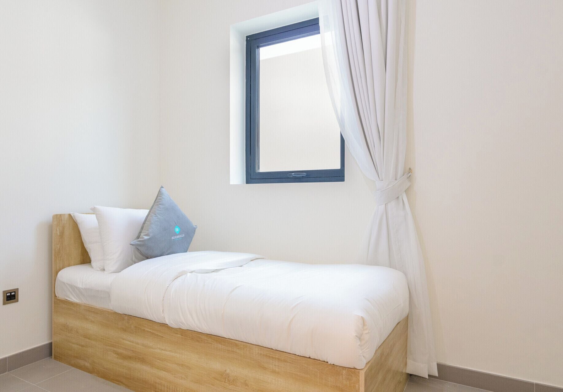 Un lit simple avec une tête de lit en bois, une literie blanche et un oreiller bleu orné d’un logo. Une fenêtre avec un cadre bleu et des rideaux blancs est visible.