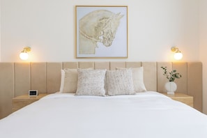A hotel room with a bed, two bedside tables, a nightstand, a framed artwork, and wall-mounted lights.