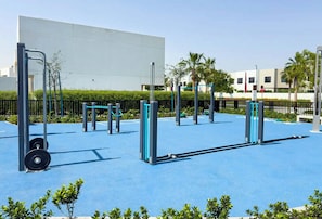 Ein Outdoor-Fitnessbereich mit Trainingsgeräten, einem blauen Gummiboden und einem weißen Gebäude im Hintergrund.