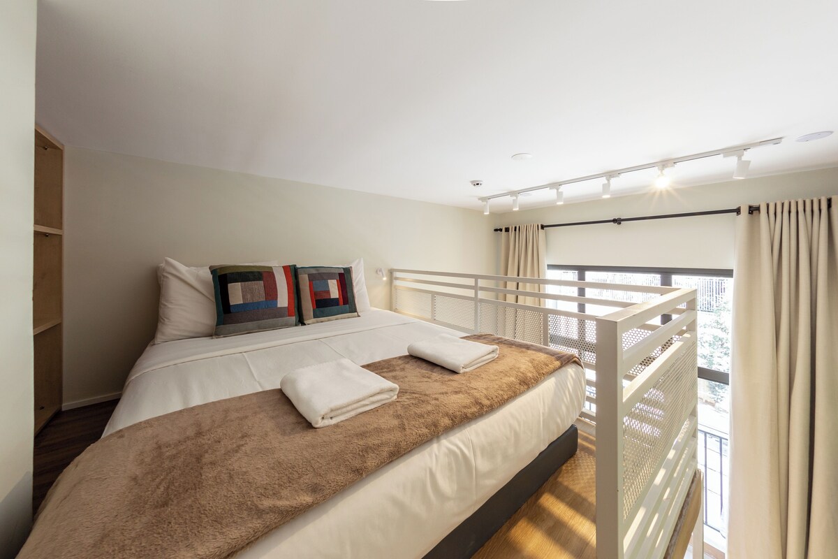 Ein Schlafzimmer mit einem Bett, Kissen, einer Decke, einem Balkon mit Geländer und Blick ins Freie.
