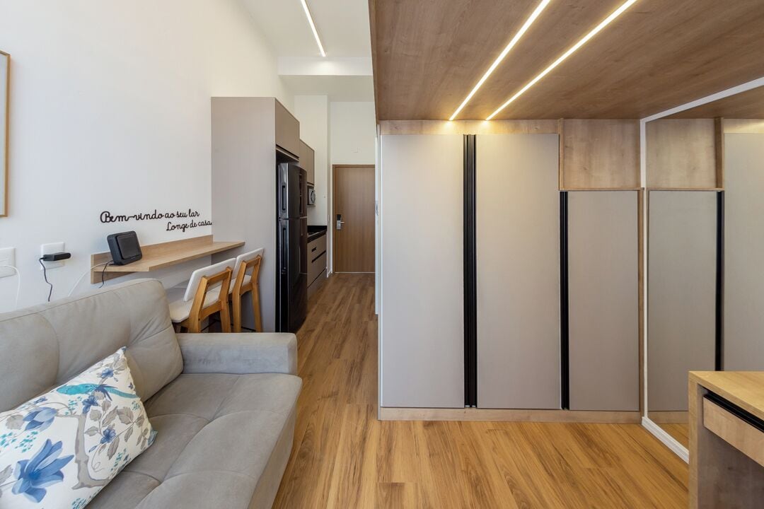 Una sala moderna con un sofá y una mesita de centro, una zona de cocina con un refrigerador y armarios, y un techo de madera con iluminación empotrada.