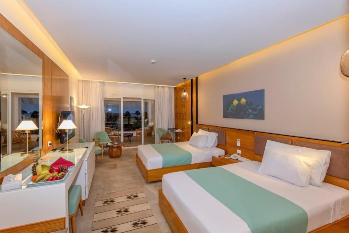 A hotel room with two beds, a desk, a chair, a dining table with fruit, and a large window with a view.