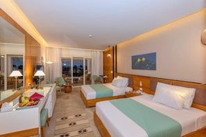 A hotel room with two beds, a desk, a chair, a dining table with fruit, and a large window with a view.