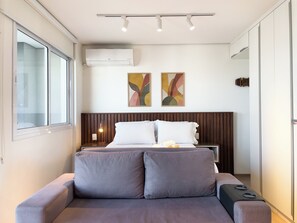 A modern bedroom with a bed, bedside table, lamp, air conditioning unit, and abstract wall art. - Ampm Paulicéia Centro (São Paulo)