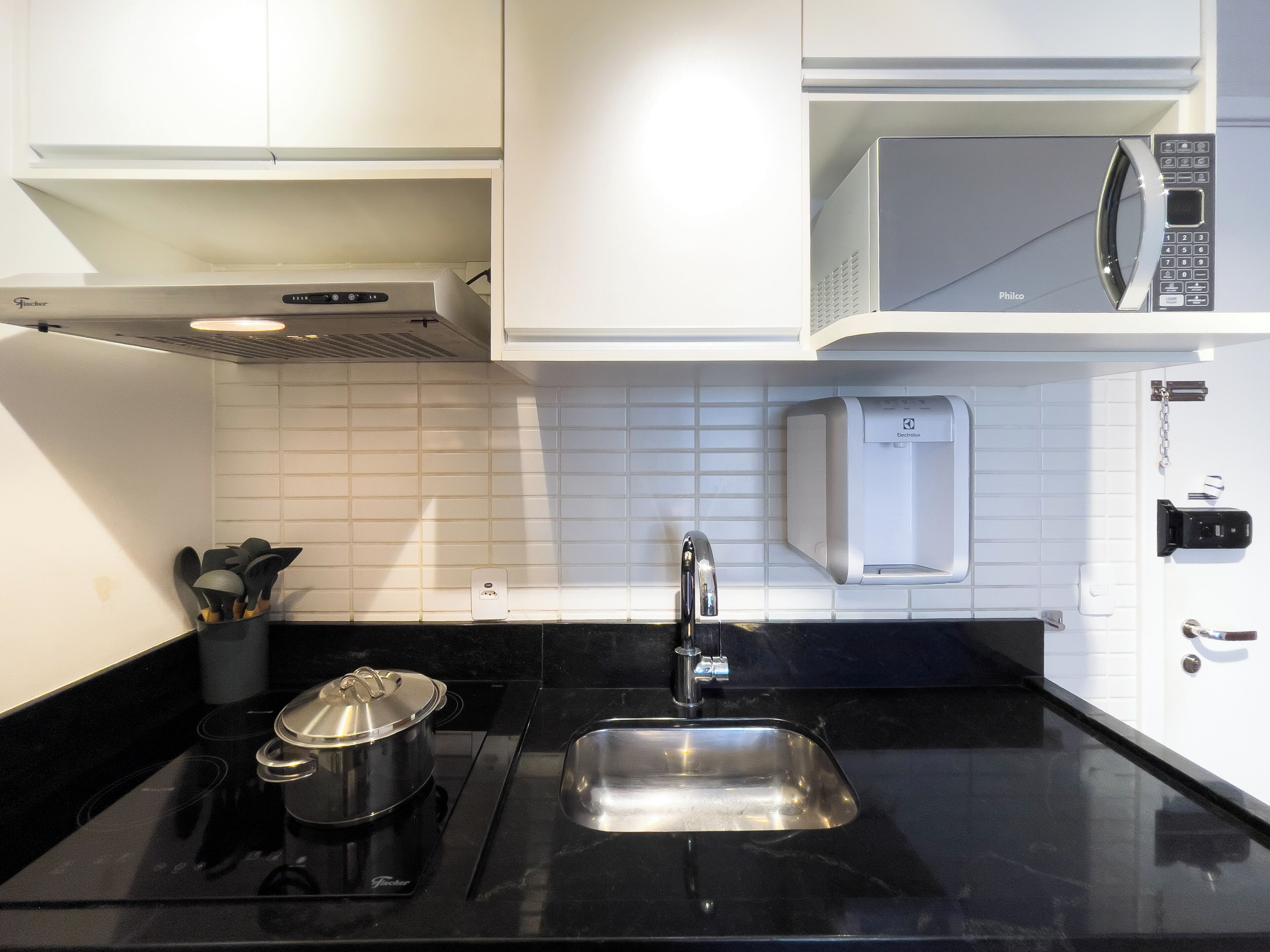 A kitchen with a black countertop, a stainless steel sink, a microwave, and a pot on the stove.
