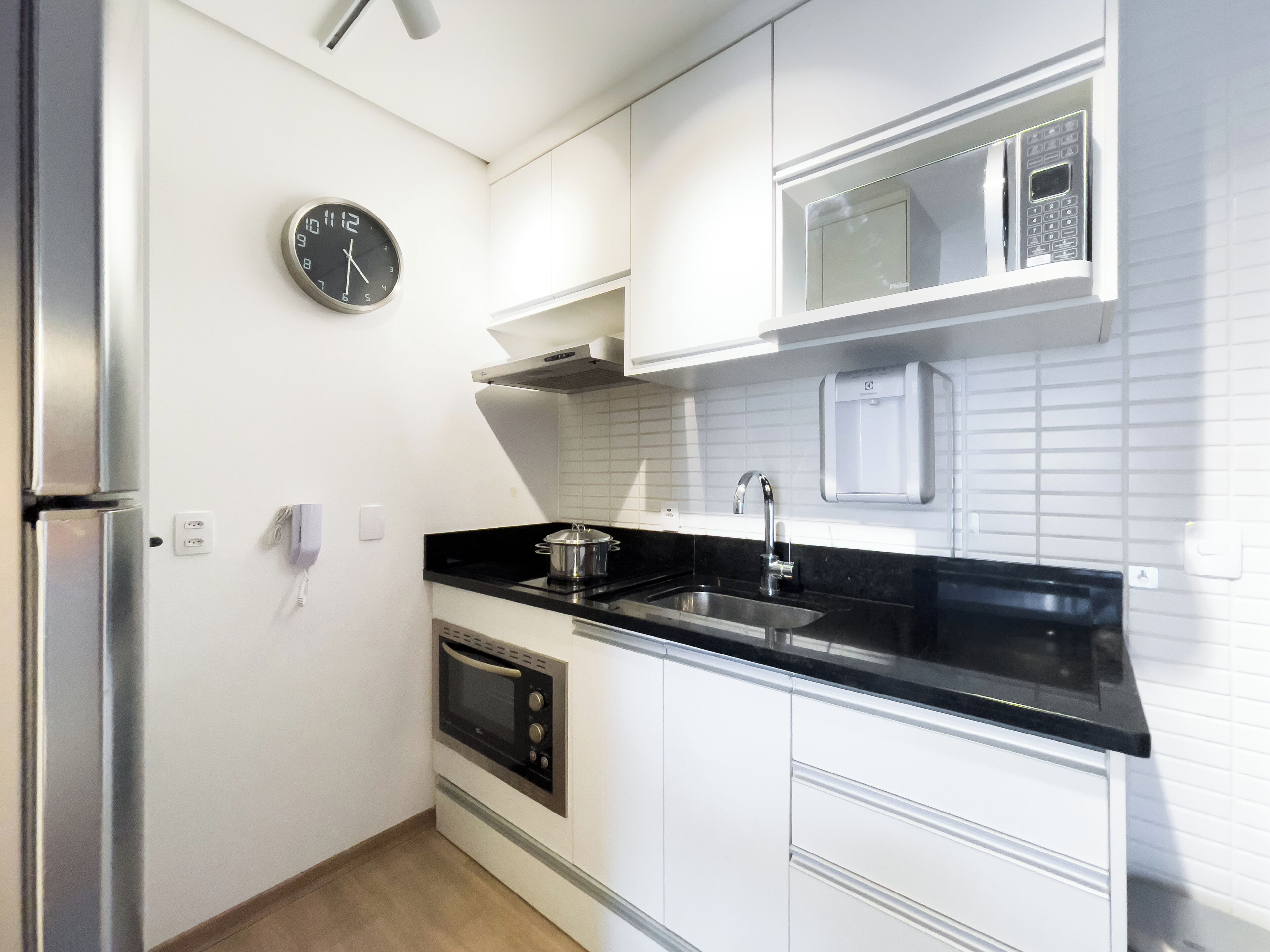 A modern kitchen with white cabinets, a black countertop, a built-in oven, a microwave, a sink, and a clock on the wall.