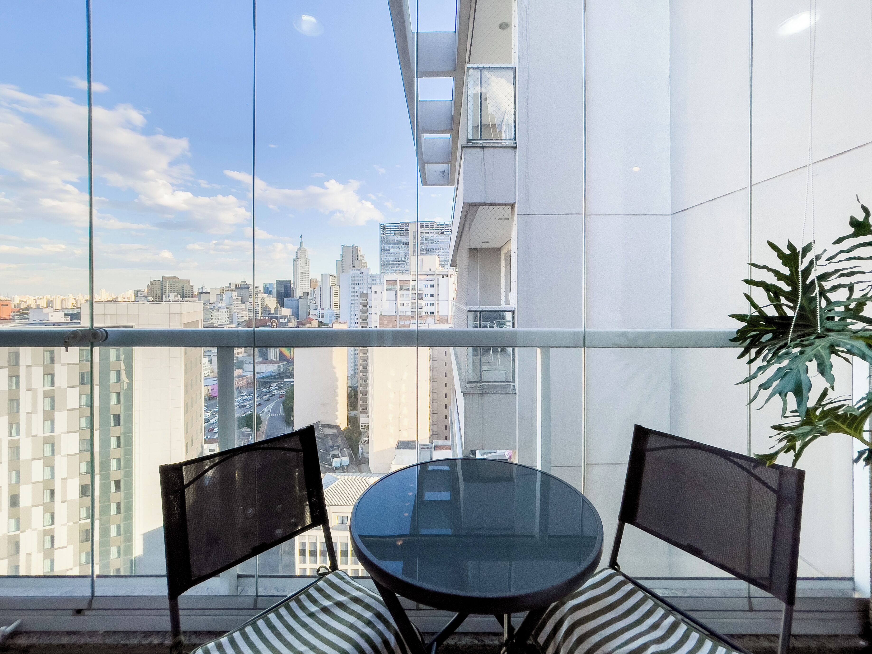 Ein Balkon mit Glasgeländer, einem runden Glastisch, zwei Stühlen und einem Blumentopf.
