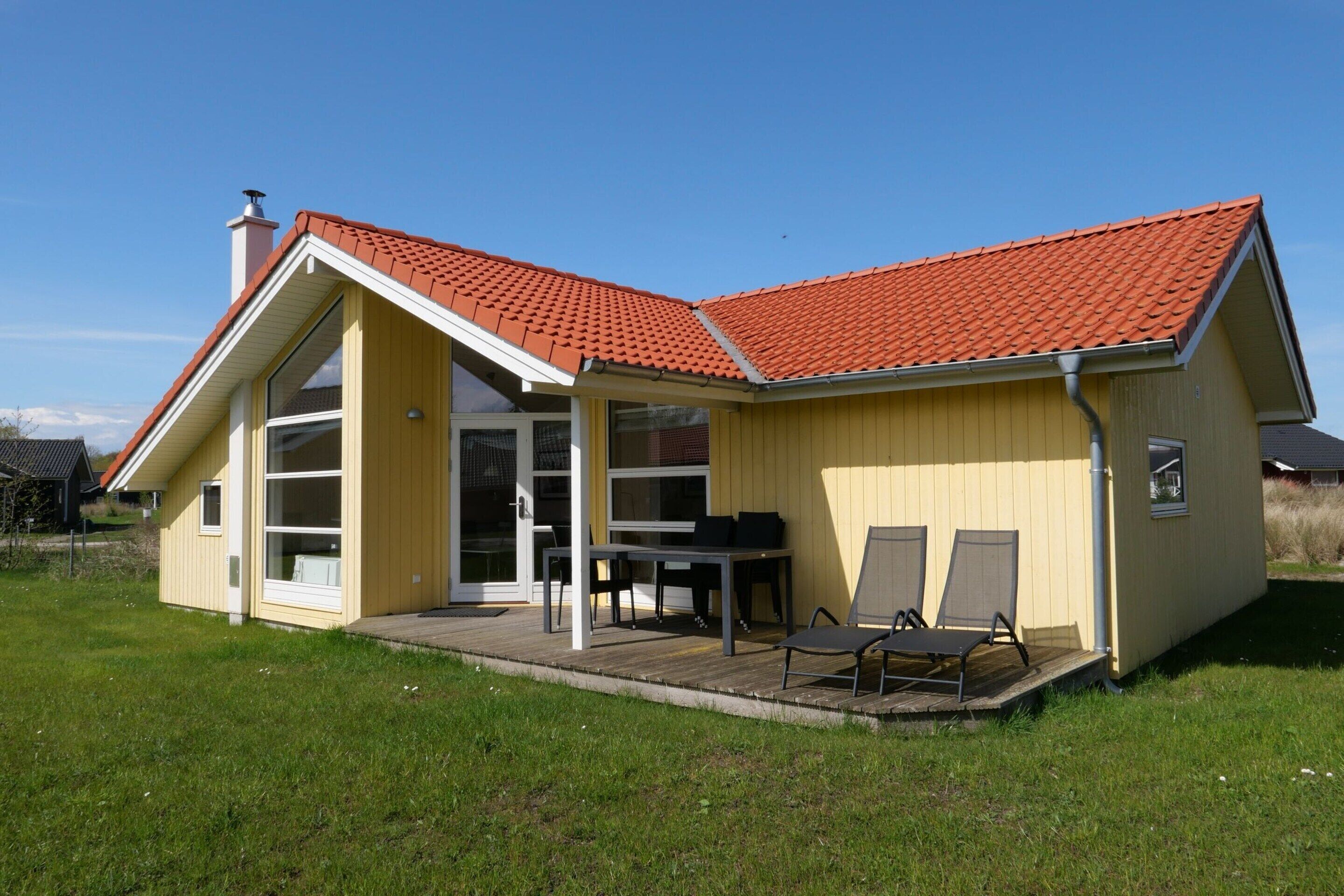 Ein eingeschossiges Haus mit rotem Ziegeldach, einer überdachten Terrasse mit Tisch und Stühlen sowie einer großen Glas-Schiebetür.