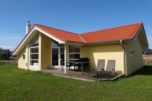 Ein eingeschossiges Haus mit rotem Ziegeldach, einer überdachten Terrasse mit Tisch und Stühlen sowie einer großen Glas-Schiebetür.